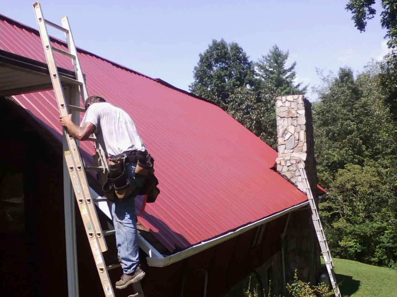 Roofer on a ladder beside a red metal roof and stone chimney — Soffit & Fascia Repair by Roofing Brothers in Saraland